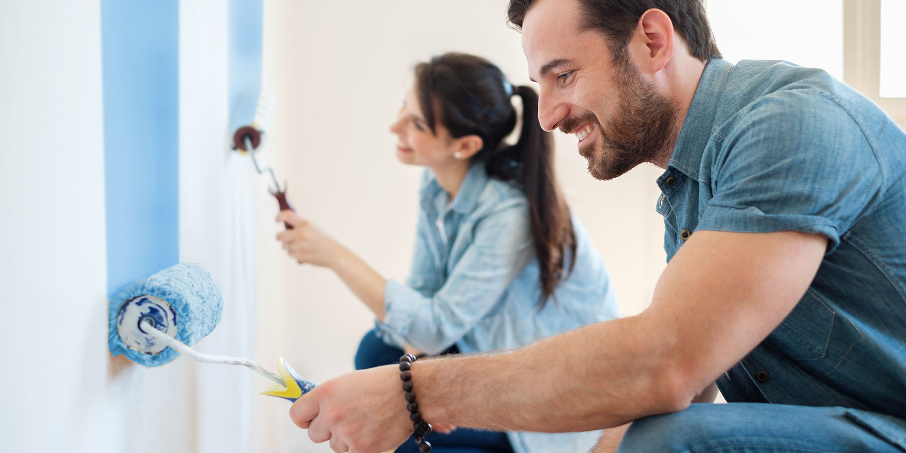 A man and woman using paint rollers to paint a wall light blue.