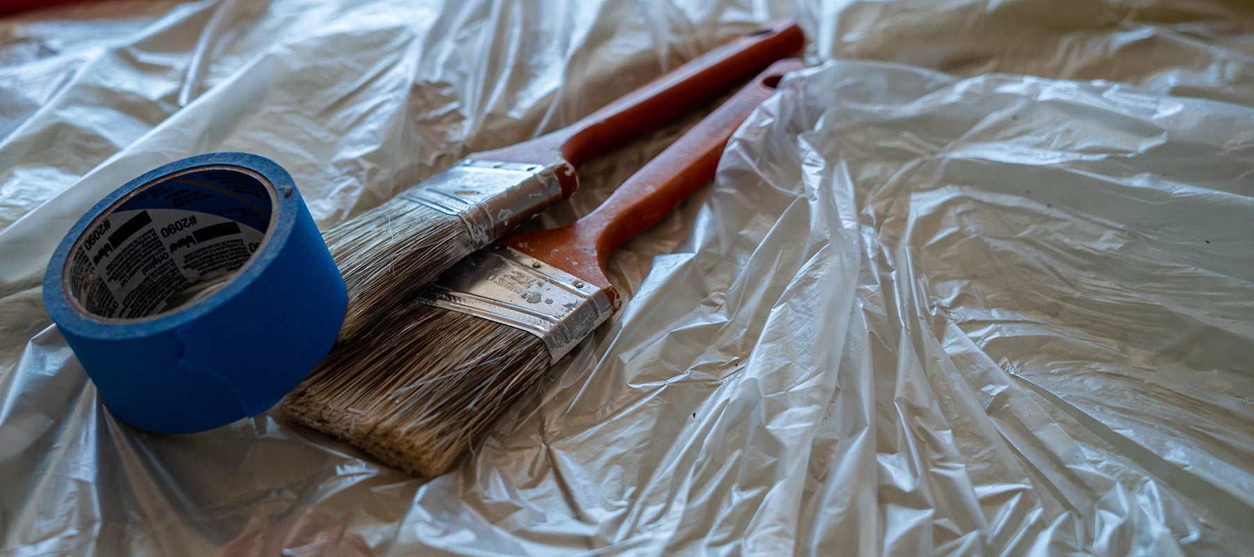 Painter's tape and two paint brushes placed on top of a plastic drop cloth.