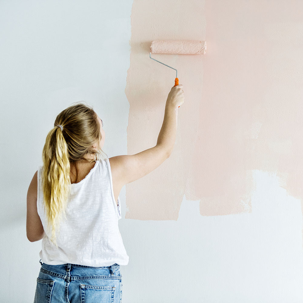 A woman painting a white wall light pink with a paint roller.